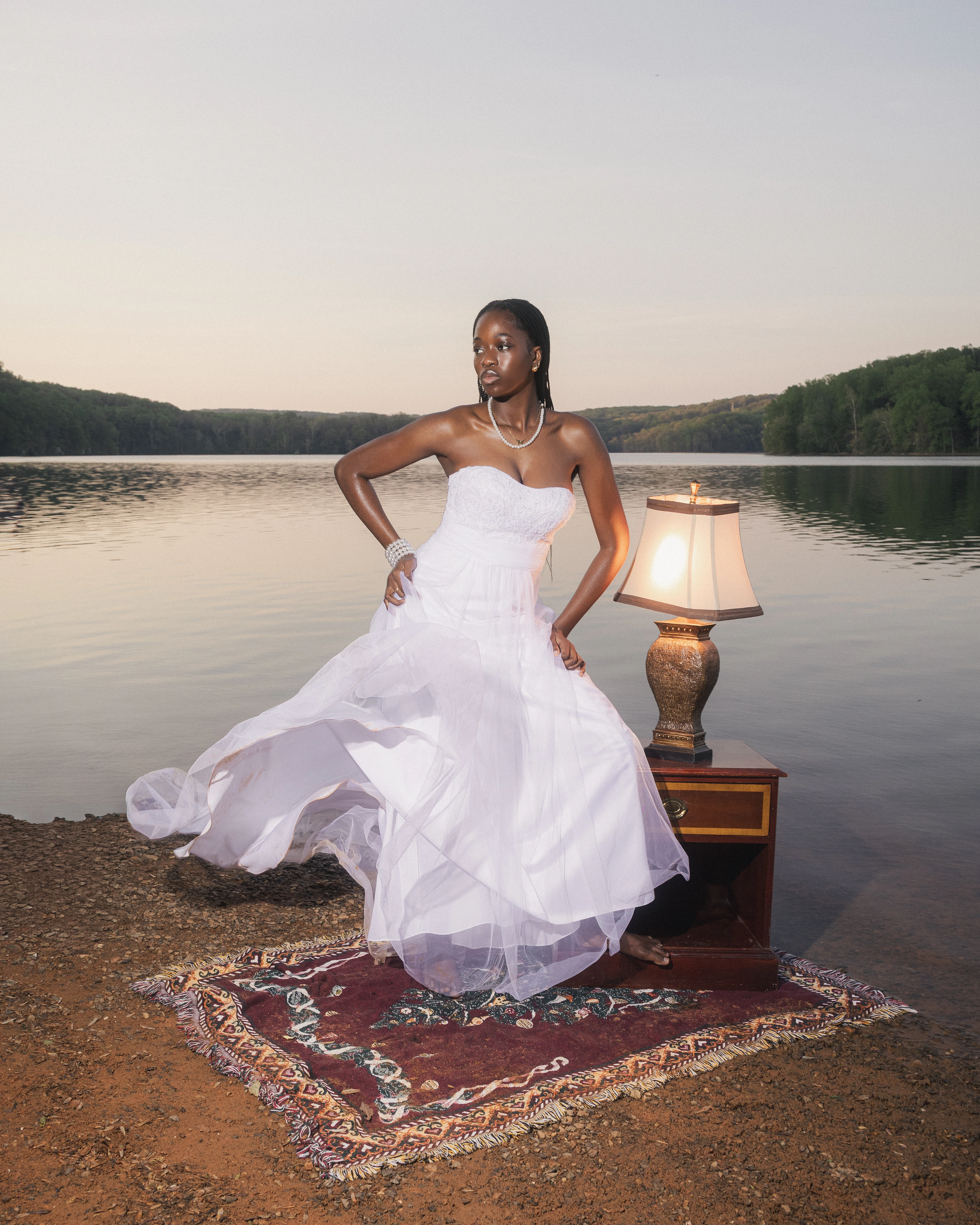 STILL editorial — wide riverside frame of model resting against an antique nightstand at dusk