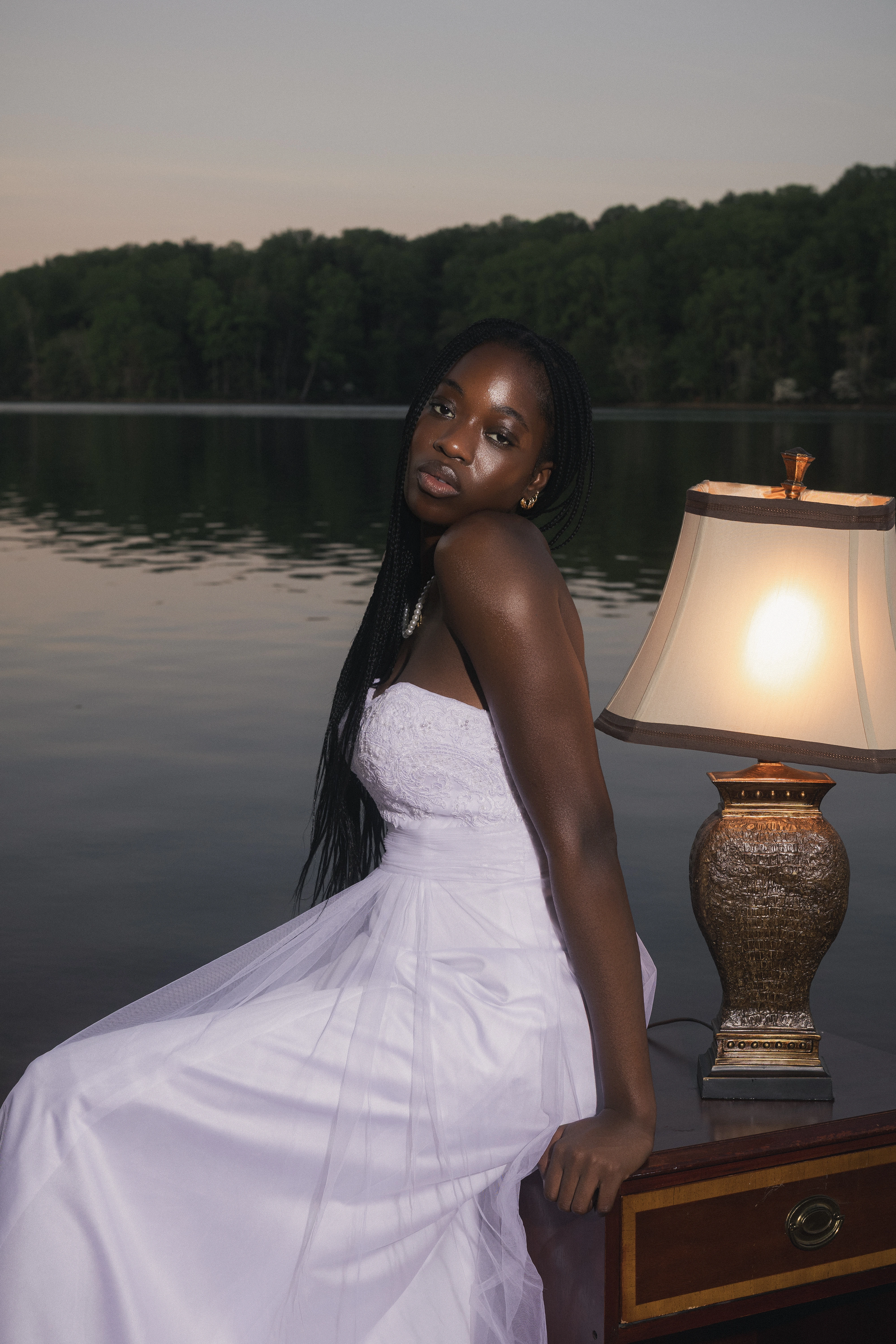 STILL editorial — model seated on a vintage chair beside a lit lamp at the lake edge